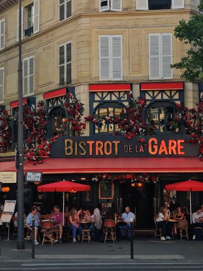Puerta del Bistrot de la Gare. Las ventanas del primer piso están cubiertas de flores rojas y debajo se ve el letrero del restaurante, con algunas luces rotas. En la puerta del restaurante hay mesas con gente cenando.