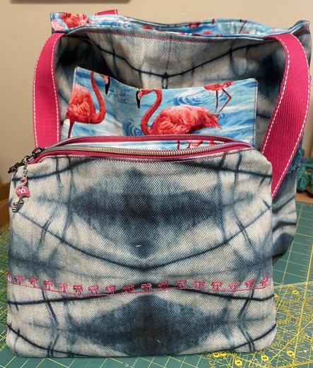 A zippered pouch in front of flamingo bag described in previous toot. The pouch has a pink zipper tape with iridescent rainbow teeth.  You can see a pink flamingo charm hanging on the left side of the open zipper. The denim has a line of pink palm trees stitched across the front. Inside, you can barely see more of the flamingo fabric.