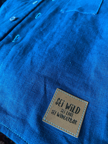 Petrolfarbenes Leinenhemd mit einem Kunstlederpatch, auf dem steht: „sei wild, seid laut, sei wunderbar“.