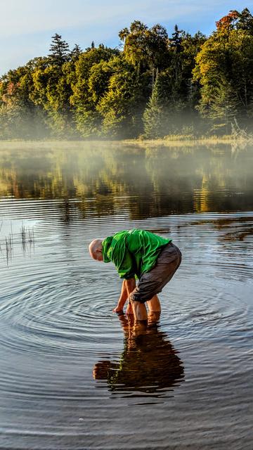 A lake sparkles in bright early morning sunshine beneath a soft blue sky. A thin mist hovers over the water near a wooded shoreline. In the foreground a man in gray shorts and a bright green hoody is bent over in water up to mid-calf, holding his water bag below the surface. Rings of concentric ripples encircle him in the otherwise placid waters.
