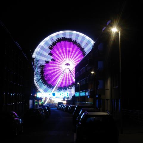 The colorful lights of a Ferris wheel form a giant pink and pzrple heart.
The Ferris wheel is framed by the black night sky and a small street with several parked cars and buildings of no more than three floors, which have a black background and shimmer blue and purple.