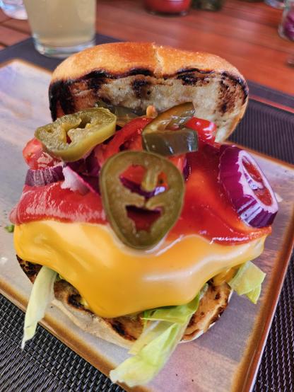 Das Bild zeigt zwei verschiedene Ansichten eines Burgers. Oben ist ein fertig zubereiteter Burger zu sehen, der auf einem Teller serviert wird. Der Burger besteht aus einem bunten Stapel von Zutaten: eine großzügige Menge geschmolzenes Käse, Jalapeños, Tomaten, rote Paprika, rote Zwiebeln und Salatblätter, alles auf einem Burgerbun. Das Bun hat einen leicht gebrannten Rand, was auf eine Grillung hinweist.