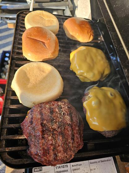 Das Bild zeigt einen Grill mit unvorbereiteten Burgerzutaten zu sehen. Ein Burgerbun wird gebraten, während ein Burgerpatty mit geschmolzenem Käse daneben liegt. Der Grill hat eine charakteristische rauchige Atmosphäre, was auf die Hitze und den Grillprozess hinweist. Der Burgerpatty hat deutliche Grillmarken, was auf eine gutes Bratenergebnis hinweist.