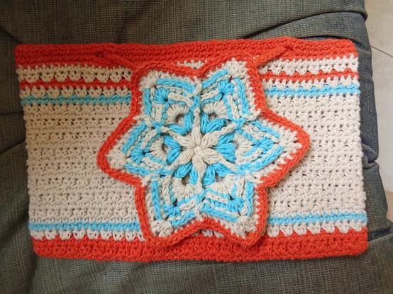 Forro rectangular de tablet tejido a crochet en una combinación de colores naranja, azul claro y blanco con una estrella grande tejida con relieves como broche en el centro y un cordón que la rodea.