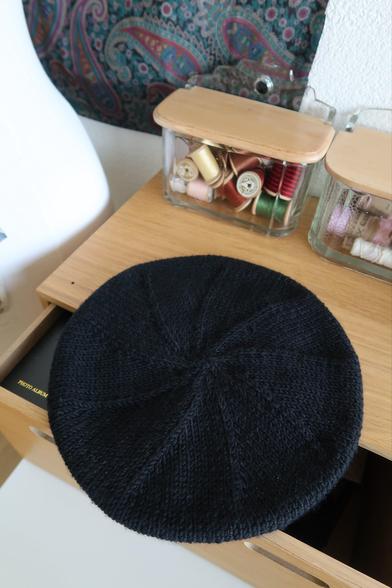 Béret noir vu d’en haut avec un motif en spirale créé par les diminutions du tricot. Derrière le bonnet, il y a deux boîtes à sel remplies de bobines.