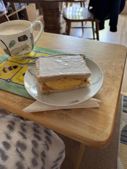 Picture a custard slice of epic proportions that fills a white side plate with a marble patttern from edge to edge! 
The plate sits next to mug of coffee, with a cat motif, on placement decorated with whimsical drawings of insects in small yellow squares with a broad green border. This in turn sits on a small wooden table.