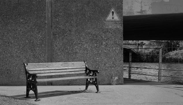 A wooden bench in front of a concrete wall and a bridge over a river to the right, the wall has a triangular exclamation mark sign, black and white image.