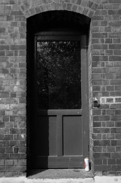 A narrow wooden door set into a wall with quite the shadow across it, the image is black and white except a can of Stella sat on the right of the doorstep.