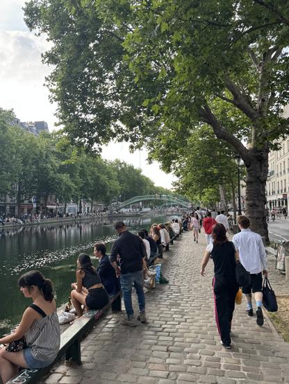 The image depicts a lively riverside scene in an urban setting, in Paris, characterized by a cobblestone pathway lined with benches and trees. In the foreground, several people are seated on the benches, enjoying the view of the calm river, while others are walking along the path. The river reflects the greenery and the sky, with a green pedestrian bridge visible in the background. The trees are lush and full, providing shade to the area. The buildings on the right side of the image are typical of Parisian architecture, with white facades and balconies. The sky is partly cloudy, suggesting a pleasant day. The overall atmosphere is relaxed and social, with people engaging in leisurely activities.
