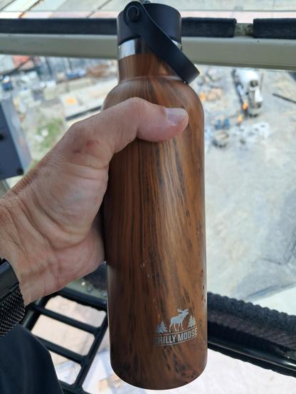 A hand holding a brown water bottle before the blurry background of a construcrion site from above.