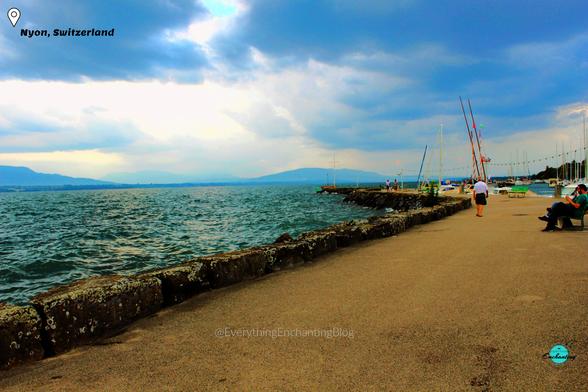 A scenic view of Geneva Lake in Nyon, Switzerland, with sailboats docked, calm waters, and a partly cloudy sky, captured from the stone-paved lakeside promenade, captured by travel blogger Anamika Chattopadhyaya for her Everything Enchanting blog and SM pages.