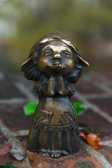 The image captures a bronze statue of a young girl in mid-prayer. She is dressed in traditional attire that includes a long skirt adorned with various patterns and pockets, indicative of historical or cultural significance. The details on her clothing are meticulously crafted, showcasing the intricate designs on both sides of her garment.

Her hands are folded together near her face, suggesting she's engaged in prayer or deep thought. She wears a hat from under which 2 long pointy ears are protruding.

The statue stands on what appears to be a brick surface, possibly indicating an outdoor setting such as a park or courtyard.