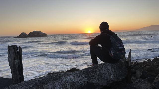 Silhouetted man sitting on a rock and looking out to sea as the sun dips below the horizon.