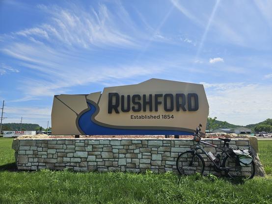 Black letters spell "Rushford established 1854" on yellow concrete sign. A blue swoop underneath it represents a river. A bicycle with a white bag leans against the grey stone wall supporting the sign. The sky has wispy white clouds over green ground.