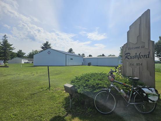 White letters spell " Welcome to Rushford est. 1854 on concrete sign of abstract design. A bicycle with a white bag leans against a short wall obscured by a bush. The grass is green under a blue sky with puffy clouds. Two white buildings for an "L" shape in the background