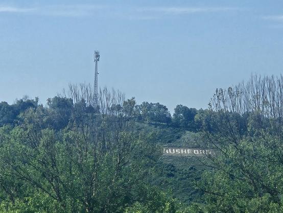 White letters spell "Rushford" on a green with bushes and trees bluff. A mobile phone tower on top of it in blue Grey sky.