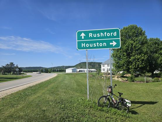 White letters spell "Rushford" with a white arrow pointing straight up, and "Houston" with a white arrow pointing to the right on a green sign. A bicycle with a white bag leans against it. The grass is green. A couple if buildings and a couple of green trees are in the background beneath a blue sky with a white cloud. To the left is an asphalt road, a white pickup truck in the distance.