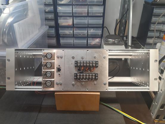 A 19" rack on a working bench with one module mounted in it. The module has a shiny front plate with 4 xlr connectors vertically arranged on the left, an ethernet connector next to it, and two rows of screw terminals on the relatively large space that makes up 2/3 of the surface on the right.