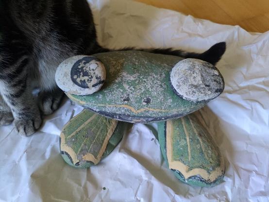 Steinfigur aus 5 Steinen. Frosch, mit 2 Froschschenkeln, einem Kopf und zwei Augen. Schwimmhäute, Nase, Mund, Augen sind grob aufgemalt. De Farbe bröselt ab, nach Jahrzehnten im Garten. Weiß, schwarz, grün, gelb-beige, rot. Unter dem Steinfrosch ist Packpapier. Links neben dem Frosch sieht man 3 Pfoten, Körper, Schwanz von unserem Tigerkater Zuro