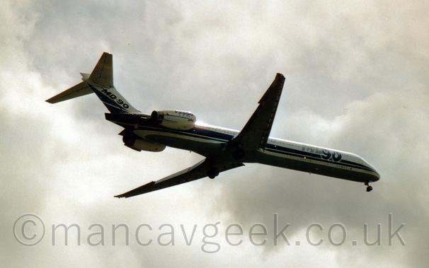 Low side view of a twin engined jet airliner flying from left to right at a very low altitude, with flaps extended from the front and rear of the wings, and undercarriage lowered, suggesting it is about to land.
The plane is mostly white, with a pair of blue stripes running along the body and a grey belly.
There are large, grey and blue "MD90" titles on the forward fuselage, with similar blue titles on the tail.
There is blue text "V2500" on the white engine pods mounted on the sides of the rear fuselage, with smaller green "IAE" titles at the front of the engine.
Somewhat dramatic grey sky fills the rest of the frame.