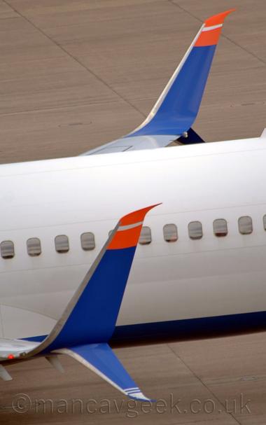 Close-up of the up-turned wing-tip of a twin engined jet airliner taxiing from right to left.
The plane is white, with a blue belly.
The tips of the wings curve upwards, painted the same blue, with the top section painted a  bright red with a white stripe.
A smaller section also curves downwards, with white stripes at the tip.
Grey concrete apron fills the rest of the frame.