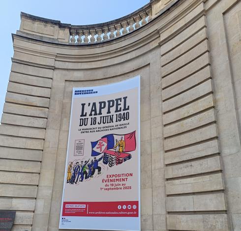 Photographie numérique couleur en format carré.
Sur un mur arrondi en pierre claire de style classique, une grande affiche présente l'exposition sur l'appel du 18 juin 1940. L'affiche est dans des tons rose, bleu, blanc et bleu, avec le dessin de 2 drapeaux (français et avec la croix de Lorraine), des personnes en train d'avancer vers de Gaulle qui lance son appel derrière un micro.