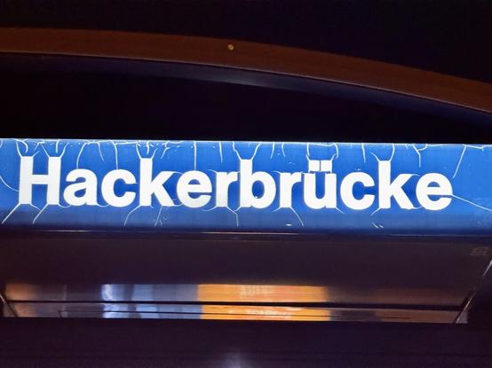 Blaues Stationsschild mit weißer Schrift: „Hackerbrücke“. Das Schild ist leicht rissig und nachts beleuchtet.
