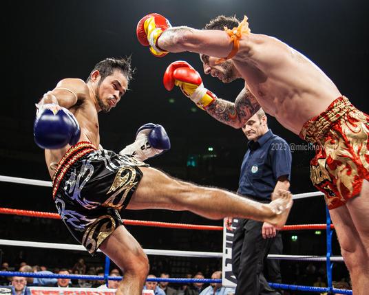 Two Muay Thai boxers. The boxer on the left, Anuwat, wearing black shorts with red and gold trim, and blue gloves is throwing a hard kick with a grimmace on his face to his opponent, Liam Harrison, who is wearing red and gold shorts and red gloves. Liam has shuffled both of his keet away from Anuwat's kick which is missing it's target - liam has his hands controlled in the air looking at Anuwat. The referee can be seen in the background looking at the action, although it looks like he blinked when the picture was taken so has his eyes closed. The referee is wearing black pants and a dark blue shirt. We can see the ropes of the boxing ring in the background and a bright spot at the top of the picture where the bright stadium lights are shinning in and lighting the action.