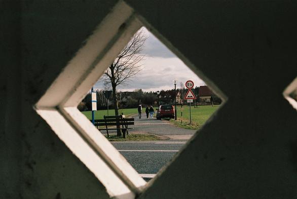 [english below] Das Foto zeigt 3 Menschen, welche eine Straße entlang laufen. Die Menschen laufen in Richtung Bildhintergrund wo Häuser stehen. Das Foto wurde durch ein kleines Fenster (ohne Glas) einer älteren Bushaltestelle aufgenommen.

[english] This photo shows three people walking down a street. The people are walking toward the background, where houses are located. The photo was taken through a small window (without glass) of an older bus stop.
