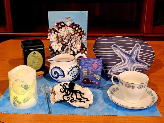 Tea tableau of an ocean scene with seashells, an octopus pendant, shells and sea stars on candle and china, paper craft, and a painted magnet. The tea is Milky Oolong loose-leaf from Harney and Sons