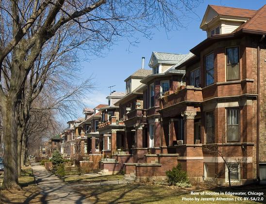 Photo of a row of two-storey, brick-built houses with dormers and basements, and with steps leading down to fairly small gardens and a footpath or sidewalk. There are trees planted between the path and the just-out-of-sight road. We are looking at the row of houses at an angle, so that the closest is on the right side of the photo, and the furthest away is closer to the left side.