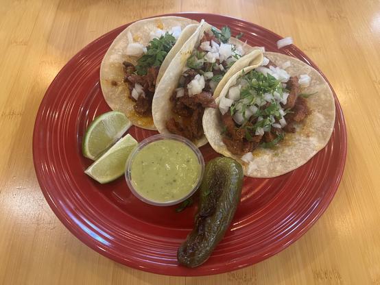 Three tacos topped with chopped onions and cilantro are on a red plate with lime wedges, a jalapeño, and a small cup of green sauce on a wooden table.