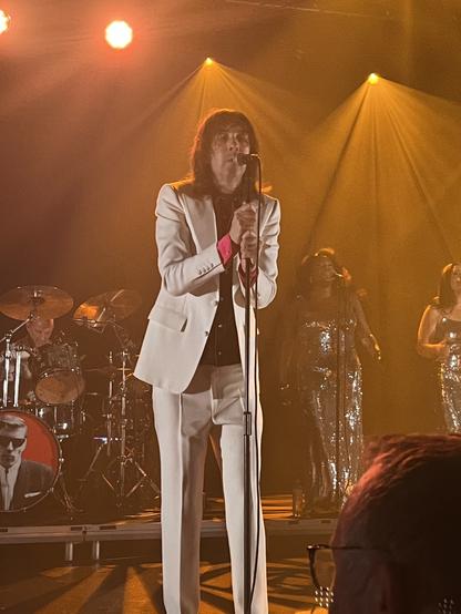 Photo of Bobbie Gillespie of Primal Scream, wearing a white suit, singing at the microphone.