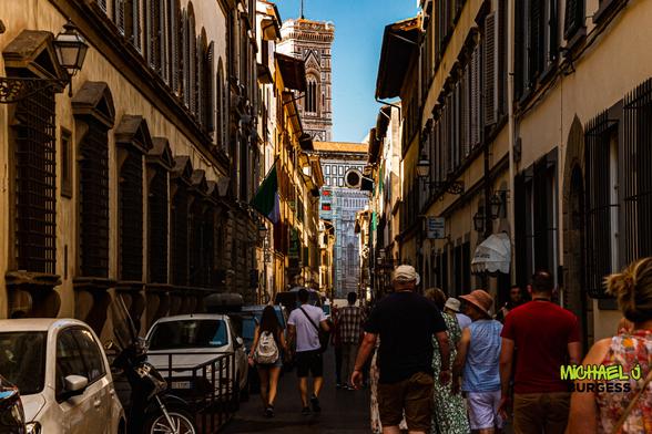 Narrow Florentine street filled with tourists walking toward the distant bell tower and façade of the Duomo; tall buildings with shuttered windows line both sides, parked cars and a scooter sit along the curb, under a clear blue sky.