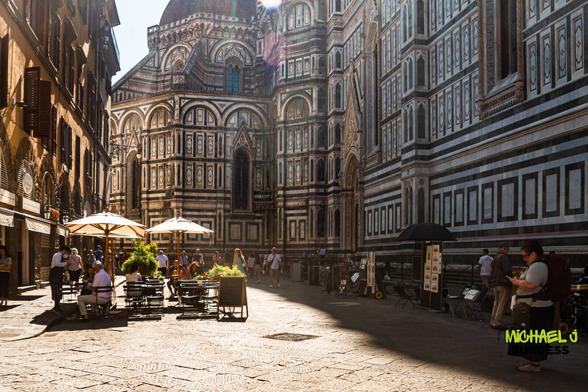 Sunlit square beside Florence’s Duomo featuring outdoor café tables with umbrellas, people chatting and walking on cobblestones, and the cathedral’s ornate multicolored marble façade rising behind.