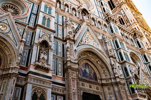 Close-up angled view of Florence Cathedral’s ornate façade showing multicolored marble panels, carved statues in niches, an arched doorway with reliefs and fresco, and part of Giotto’s bell tower rising at the edge.