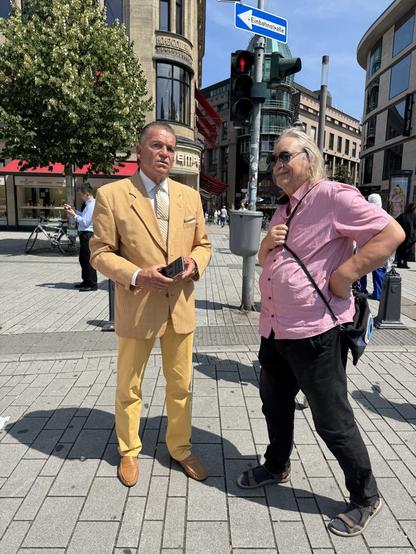 Vom Look her könnten diese beiden Herren nicht unterschiedlicher sein. Links ein Mann mit zurück gegelten Haaren und im teuren (?) gelben Maßanzug, Rechts ein Herr (Wöske) von der Mobilitätswende im legeren Kurzarmhemd mit Sonnenbrille und langen Haaren, der seine Sandalen ausführt, doch beide befürworten einen wirklich autofreien Corneliusplatz.