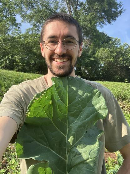 a smiling man holds a collard green up to his torso, showing that it's as big as his chest; he appears proud of this harvest from his garden
