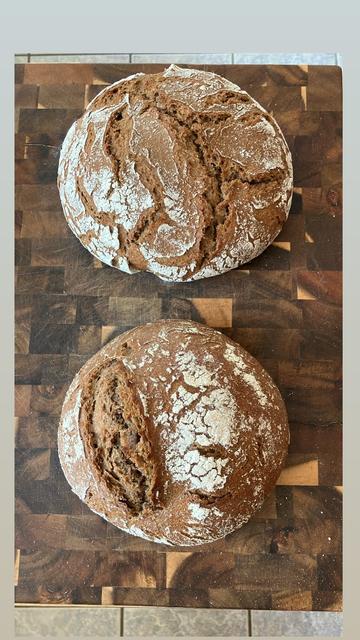two gorgeous loaves of sourdough rye bread sit atop a dark wooden cutting board; both are powdered with white rye flower, giving extra contrast to the cracks and seams, shallow and deep, that give definition to the surface of the bread