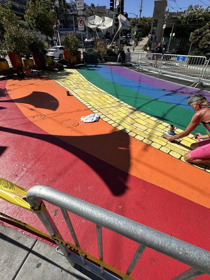 A person in a rainbow shirt paints in the “stones” of the yellow stripe on the sidewalk at Jane Warner Plaza in San Francisco’s Castro district