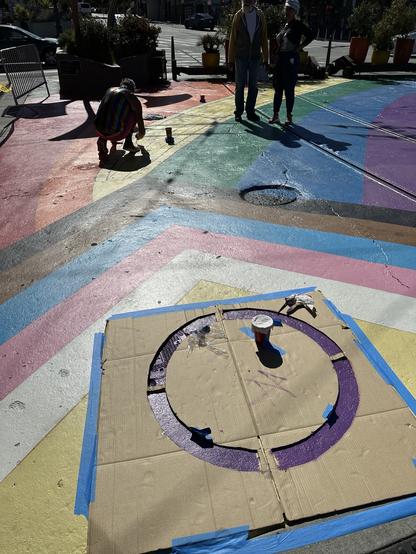 A shot of people watching final touches added to the rainbow Progress Flag being painted at Jane Warner Plaza in San Francisco. The purple circle, symbolizing Intersex people, is drying under a cardboard template in the foreground.