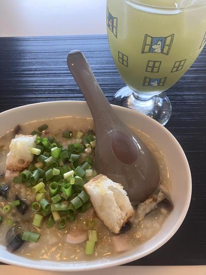 A bowl of congee topped with chopped green onions, pieces of bread, and a spoon. Next to it is a glass of green drink with a cartoon design. The background features a textured surface.