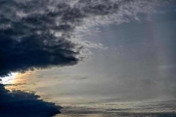 Color photo of clouds in the sky. On the left, they are thick and dark, but an angled gap reveals the bright light of the hidden sun. In the middle and the right, thin wispy clouds are present. A rainbow arc, barely visible, is on the extreme right of the frame.