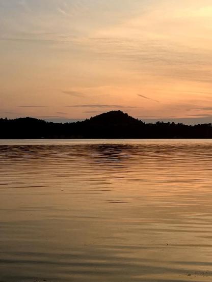 A serene lake scene at last night’s sunset, featuring calm waters reflecting hues of orange and pink, with a silhouette of a hill in the background.