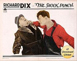 Lobby card of the film. A man is punching another man on the chin. The punching man is wearing a dark overalls and a long-sleeved red shirt. The punched man is wearing a brown coat and a gray hat.