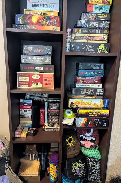 Image depicts a small brown cubbyhole style bookcase containing a multitude of small board, card and dice games.