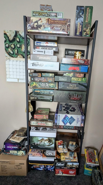 Image depicts a vertical open frame bookcase containing many board games and boxes of terrain and unpainted minis.