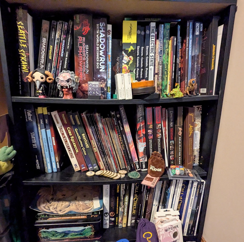Image depicts 3 shelves of a cheap black bookcase groaning under the weight of many Role Playing Game sourcebooks, including Shadowrun, Cyberpunk, Bunkers & Badasses, Fallout, Pathfinder, DragonAGE, Firefly, Call of Cthulhu and many more.