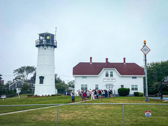 A group of people is gathered in front of a white U.S. Coast Guard building with a red roof. To the left stands a tall, white lighthouse with a black railing around the top, its light faintly visible through the glass. The building and lighthouse sit on a neatly trimmed lawn surrounded by a low fence. On the far right, a tall warning pole has a blinking yellow light and a diamond-shaped sign that reads “DANGER.” The sky is gray and overcast, and trees and houses are visible in the background.