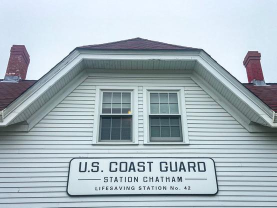 The image shows the upper part of a white wooden building with a steep roof and two red brick chimneys. Below a pair of double windows is a large rectangular sign that reads: "U.S. COAST GUARD – Station Chatham – Lifesaving Station No. 42." The sky in the background is overcast.